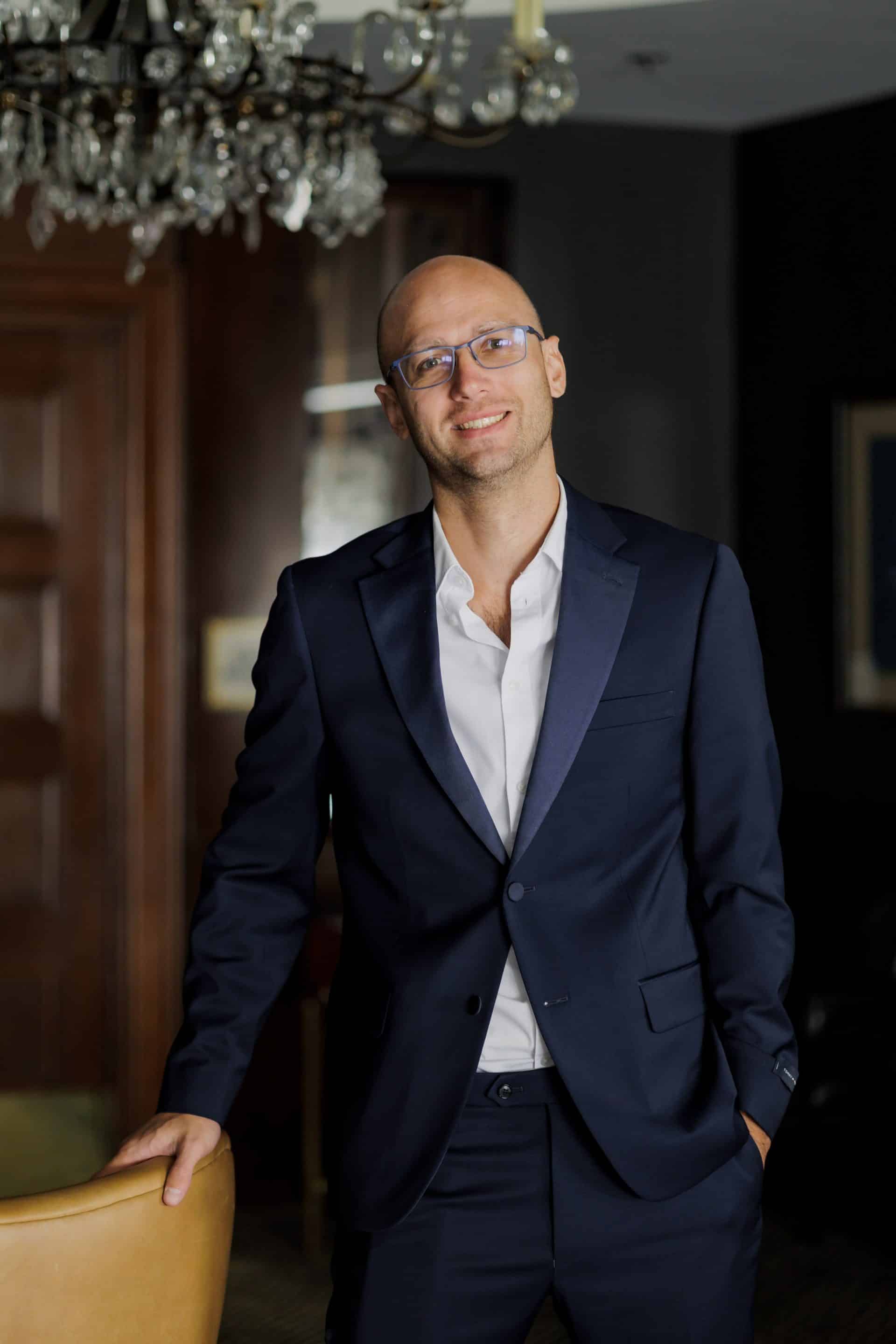 A man with glasses, dressed in a dark blue suit and white shirt, stands smiling with one hand on a chair in a dimly lit room. The background includes a wooden door, a chandelier, and a picture frame.