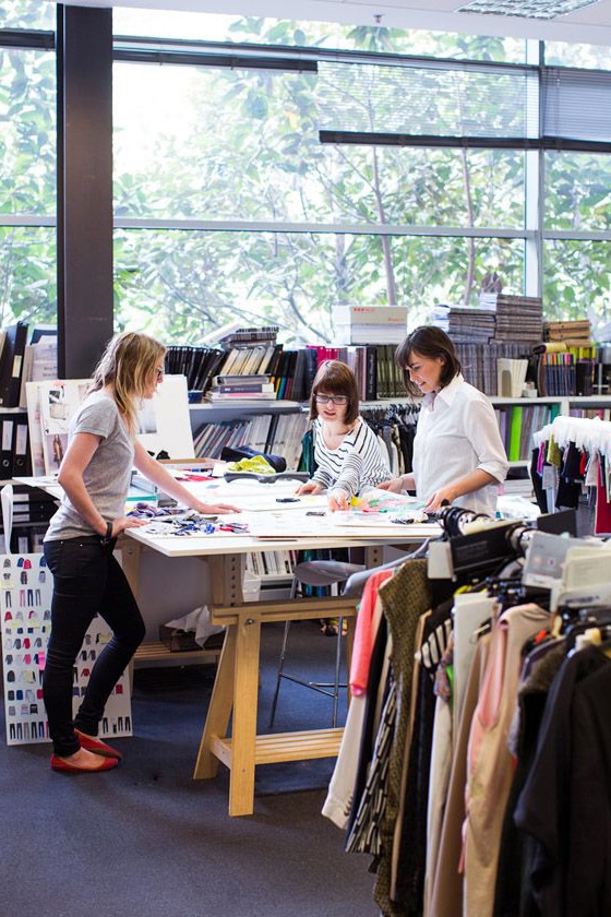 Three women are working at a large table in a well-lit room filled with fabrics, books, and garments. The table is scattered with various materials and design tools. Clothing racks with different garments surround them, indicating a creative workspace.