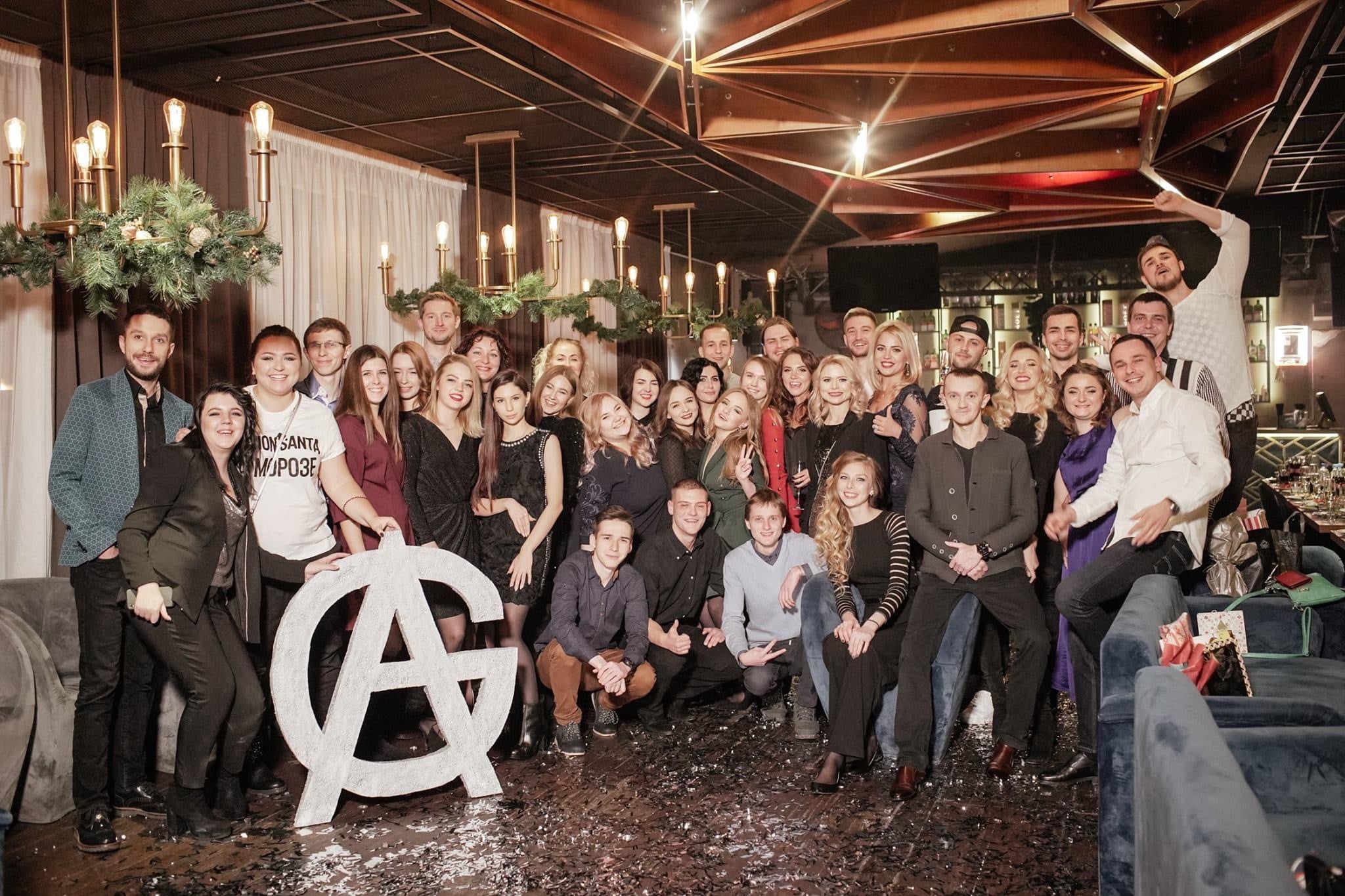 A large group of people at a festive indoor event pose for a group photo. Some hold drinks and others smile at the camera. There's greenery decor and ambient lighting in the background, with a big decorative "A" behind the group. Confetti covers the floor.