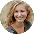 A young woman with long, wavy blonde hair is smiling and looking at the camera. She is wearing a green top and a delicate necklace. Behind her, there is a blurred background of tall grass and natural outdoor scenery.